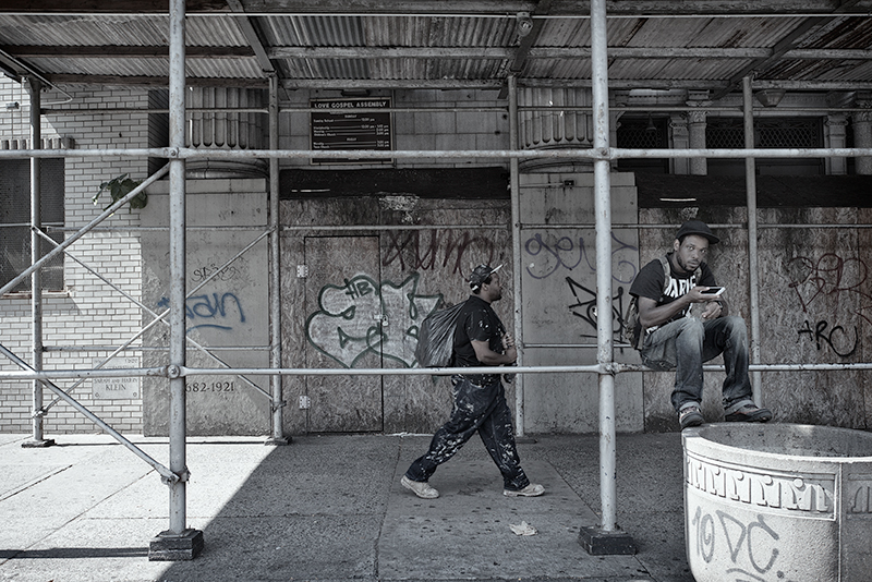 Young man sits on scaffolding in front of old graffitied building, while another passes by.