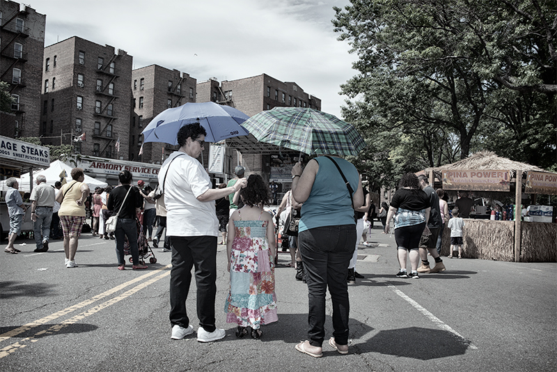 Women and little girl walking under umbrellas; hot day at a street fair.