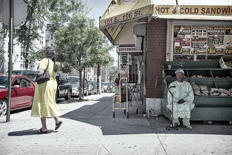 Man seated in shade at supermarket; woman approaches intersection.