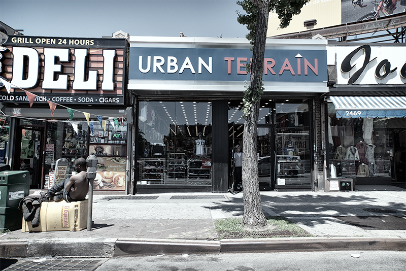 Young man seated on boxes in front of store fronts; one is a deli, one is "Urban Terrain."