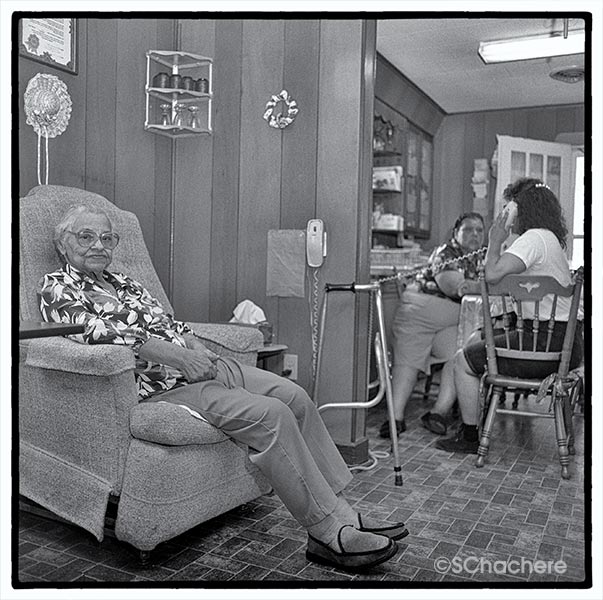 Older woman in recliner, others in kitchen in background.